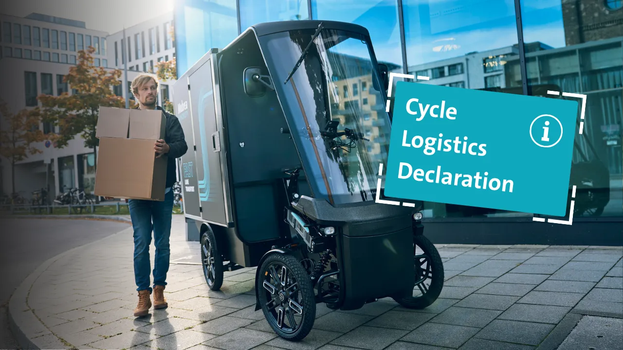 Lieferfahrer trägt Kartons neben einem Mubea Cargobike vor einem modernen Bürogebäude.