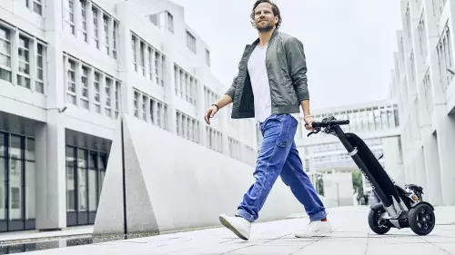 Ein Mann in lässiger Kleidung, bestehend aus Jeans, einem T-Shirt und einer Jacke, geht durch eine moderne städtische Umgebung und zieht dabei einen E-Scooter hinter sich her. Im Hintergrund sind hohe, weiße Gebäude mit vielen Fenstern zu sehen.