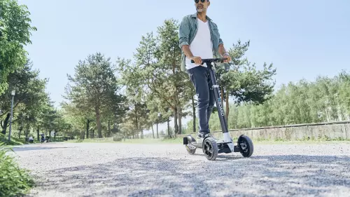 Ein Mann mit Sonnenbrille, weißem T-Shirt und Jeansjacke fährt auf einem XBoard-Elektroroller auf einem Schotterweg in einem sonnigen Park. Umgeben von hohen Bäumen genießt er die Fahrt durch die grüne Umgebung.