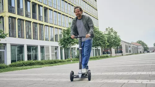 Dan (ein Mann) fährt in Freizeitkleidung auf einem XBoard in einer Stadt. Im Hintergrund sind Gebäude zu sehen.