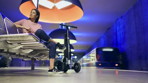 Mia (eine Frau) sitzt in Freizeitkleidung auf einer Bank in einer U-Bahn-Station. Neben ihr steht eine XBoard.