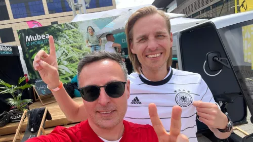  Zwei Männer lächeln und posieren in Spanien- und Deutschland-Fußballtrikots, während sie das Peace-Zeichen machen. Sie stehen vor einem Mubea U-Mobility-Stand bei einer Outdoor-Veranstaltung.
