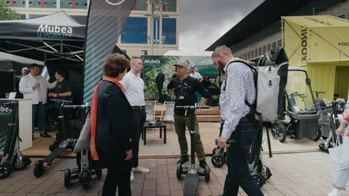 Eine Gruppe von Personen steht um den Mubea U-Mobility Stand bei einer Outdoor-Veranstaltung. Einige unterhalten sich, während andere die ausgestellten XBOARD Elektroroller und weiteren Mobilitätslösungen begutachten.