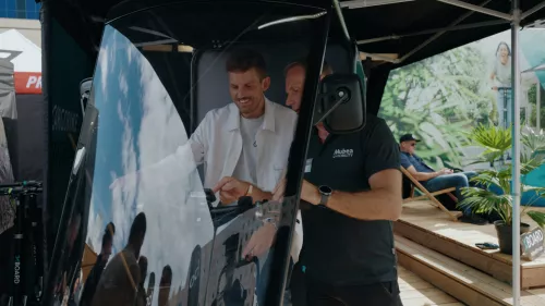 Zwei Männer stehen hinter dem Cockpit eines Cargobikes. Ein Mann erklärt die Bedienelemente, während der andere interessiert zuschaut und lächelt. Im Hintergrund ist ein Veranstaltungsstand zu sehen, und eine Person sitzt entspannt.