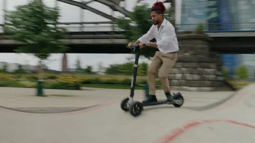 Ein Mann mit roten Dreadlocks fährt auf einem XBOARD Elektroroller durch eine städtische Umgebung. Im Hintergrund sind eine Brücke und Grünflächen zu sehen. Das Bild fängt die Bewegung ein, während er den Roller auf einem glatten Weg testet.