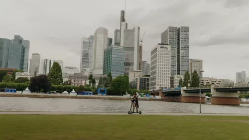 Eine Person fährt auf einem XBOARD Elektroroller entlang eines Flussufers, im Hintergrund Frankfurt mit moderne Wolkenkratzer und Gebäude. Die Szene zeigt einen ruhigen Fluss, grünes Gras und einen bewölkten Himmel. Das Stadtpanorama und eine Brücke sind deutlich im Hintergrund zu sehen.