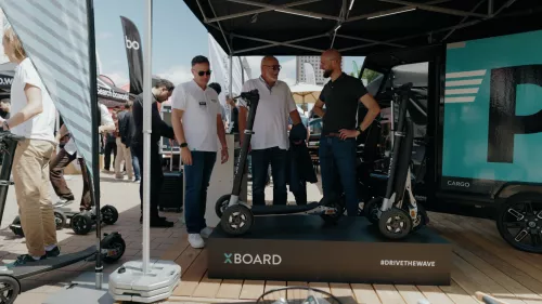Eine Gruppe Männer steht neben einem ausgestellten XBOARD Elektroroller und unterhält sich am Mubea U-Mobility Stand bei einer Outdoor-Veranstaltung. Der Stand zeigt ein Schild mit der Aufschrift „DRIVE THE WAVE“ und verschiedene elektrische Mobilitätslösungen sind ausgestellt.