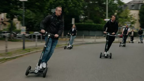 Eine Gruppe von Menschen fährt auf XBOARD Elektrorollern durch eine Stadtstraße. Sie lehnen sich leicht in die Kurven, während sie zusammen fahren. Im Hintergrund sind parkende Autos, Bäume und einige Gebäude zu sehen.