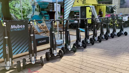  Eine Reihe von XBOARD-Elektrorollern ist ordentlich vor einem Mubea U-Mobility-Ausstellungsstand aufgereiht. Im Hintergrund sind Werbematerialien zu sehen, darunter ein Banner mit einer Person, die einen XBOARD-Roller fährt, Stühle und ein kleines Fahrzeug im Stil eines Food-Trucks. Die Szene scheint Teil einer Outdoor-Ausstellung oder eines Events zu sein.