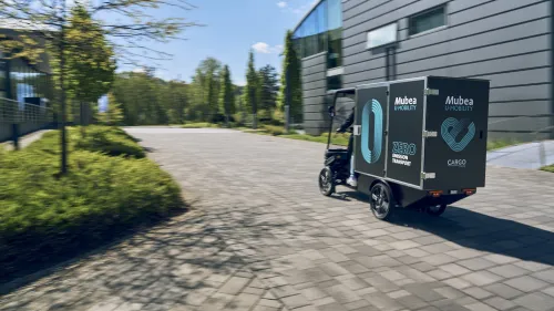 E-Cargobike mit großem Laderaum fährt auf gepflastertem Weg an modernen Gebäude entlang.