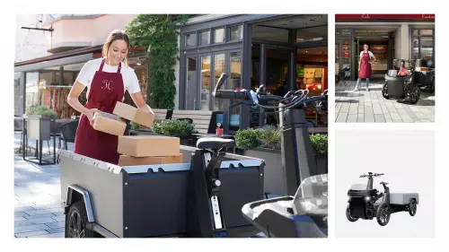 The image shows a woman working with the Mubea Cargo cargo bike in an urban setting. She is wearing a red apron and lifting several boxes from the vehicle's cargo area. In the background, shops and tables are visible, highlighting the practical use of the Cargo for deliveries and transportation in the city.
