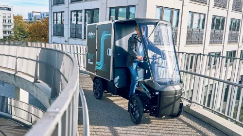 PACK fährt auf einer Fahrradbrücke vor modernen Gebäuden.