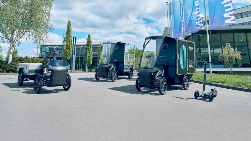 Drei E-Cargobikes vor Mubea-Gebäude mit Fahnen, Bäumen und blauem Himmel im Hintergrund.